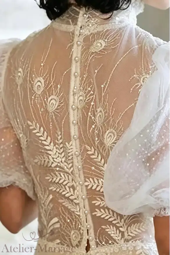Gros plan sur l'alignement délicat des boutons perles et la broderie fine sur le dos transparent d'une robe de mariée vintage à col montant.