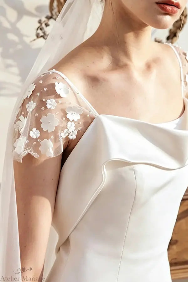 Détail délicat de l'épaule d'une robe de mariée en satin fluide, ornée de fleurs 3D artisanales et perles sur un tulle invisible.