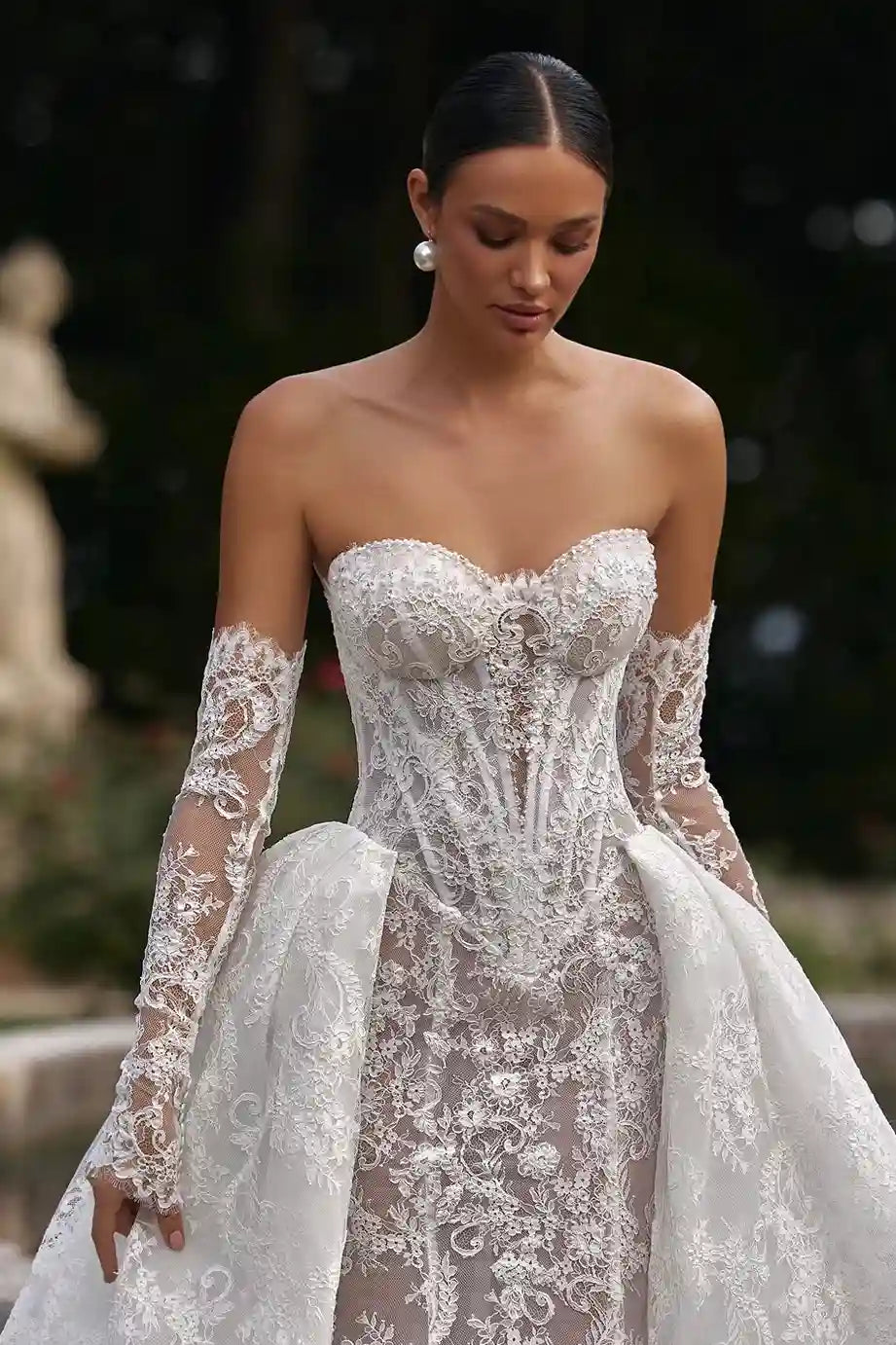 Vue de face d'une mariée près d'une fontaine, mettant en valeur la coupe ajustée et l'effet tatouage floral de ses manches amovibles.