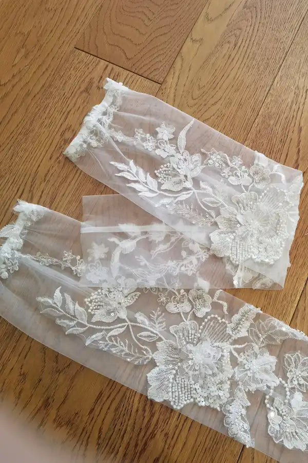Paire de manches amovibles de mariée en dentelle perlée blanche, posées à plat sur un parquet en bois, montrant la longueur et les motifs floraux délicats.