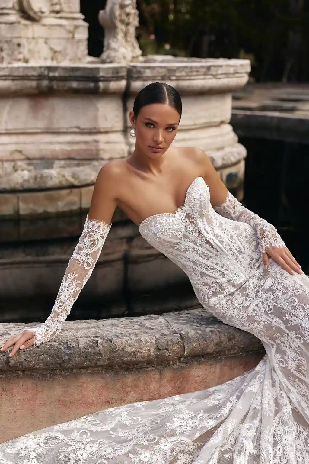 Mariée adossée à une fontaine en pierre, illustrant parfaitement la ligne épurée et non bouffante de ces manches amovibles en dentelle.