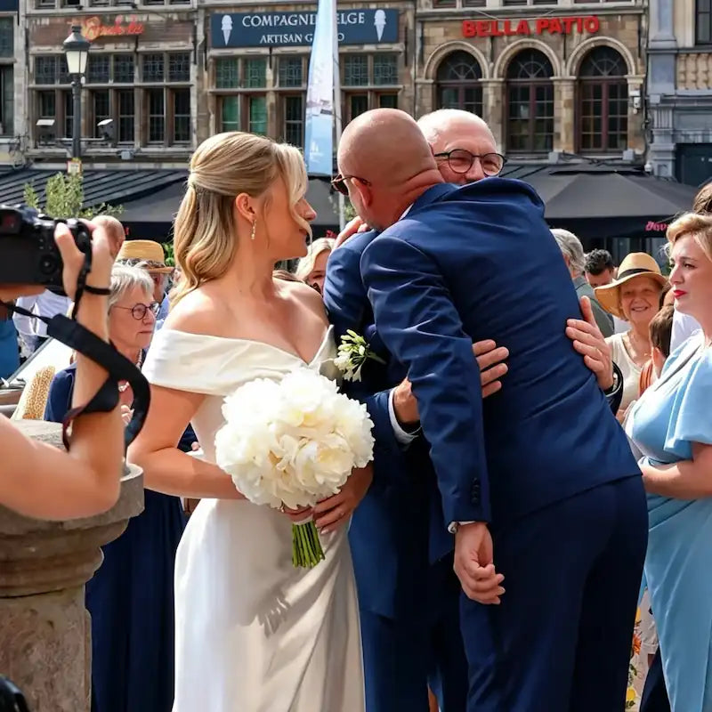 Émotion spontanée : une mariée rayonnante en robe col bardot observe une tendre étreinte entre ses proches sur la Grote Markt d'Anvers