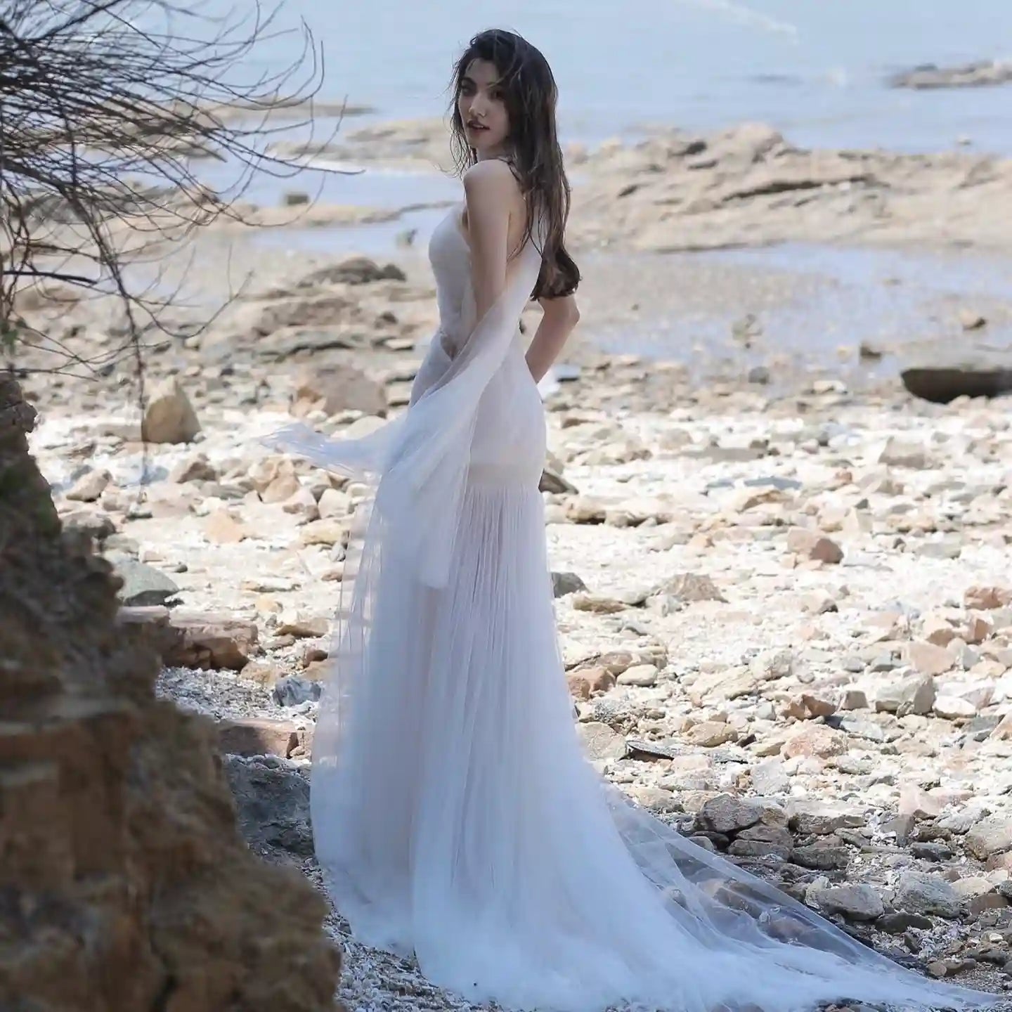 Mariée de dos sur une plage rocheuse, sa robe de mariée fluide et vaporeuse flotte délicatement au vent, illustrant la liberté de mouvement