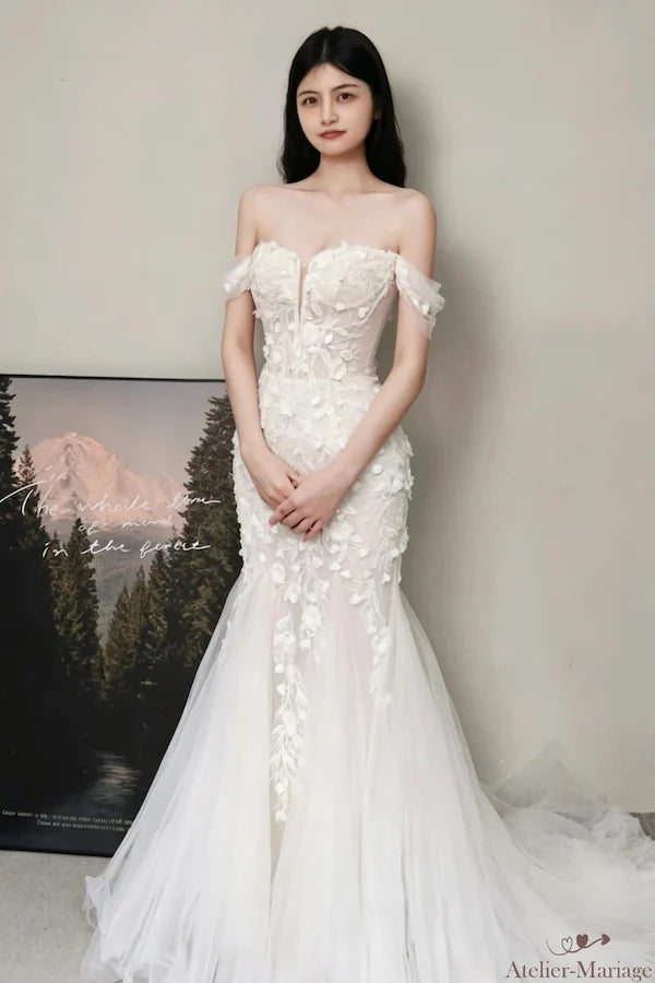 Vue complète d'une mariée portant la robe, posant élégamment dans un décor bohème chic. Silhouette sirène, manches off-shoulder et jupe en tulle. Laissez-vous séduire par cette robe de créateur.