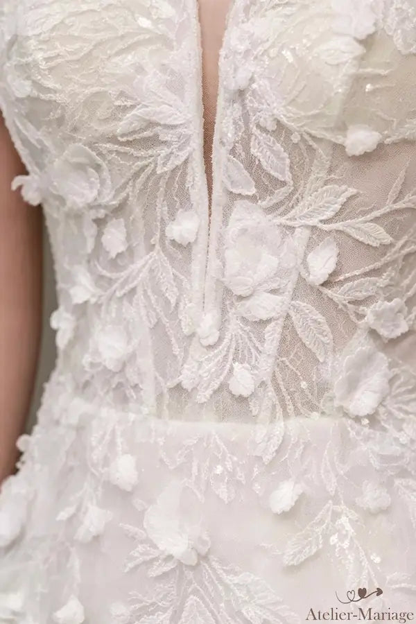 Gros plan détaillé du corsage de la robe de mariée, montrant le décolleté plongeant illusion, la dentelle blanche délicate et les perles. Signature Atelier Mariage.