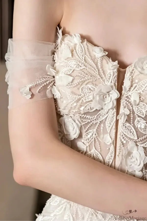 Vue rapprochée du bras et du décolleté de la robe de mariée bohème. Détails de la dentelle florale 3D et de la manche en tulle. Marque Atelier Mariage. Commandez votre robe sur mesure.