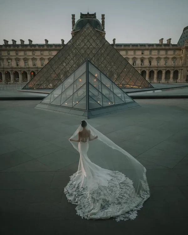 Mariée de dos devant la pyramide du Louvre portant une robe de mariée sirène romantique avec longue traîne et voile transparent