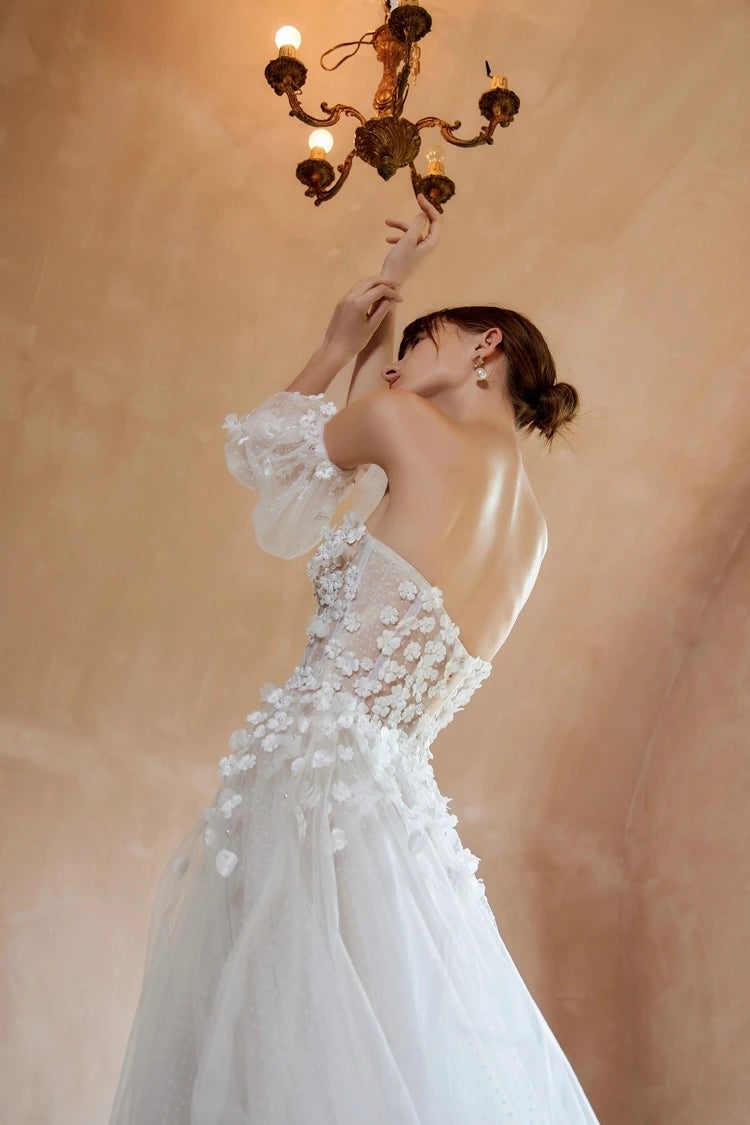 Mariée posant sous un lustre vintage, dévoilant le profil et le dos d'une robe romantique à fleurs 3D avec manches bouffantes.