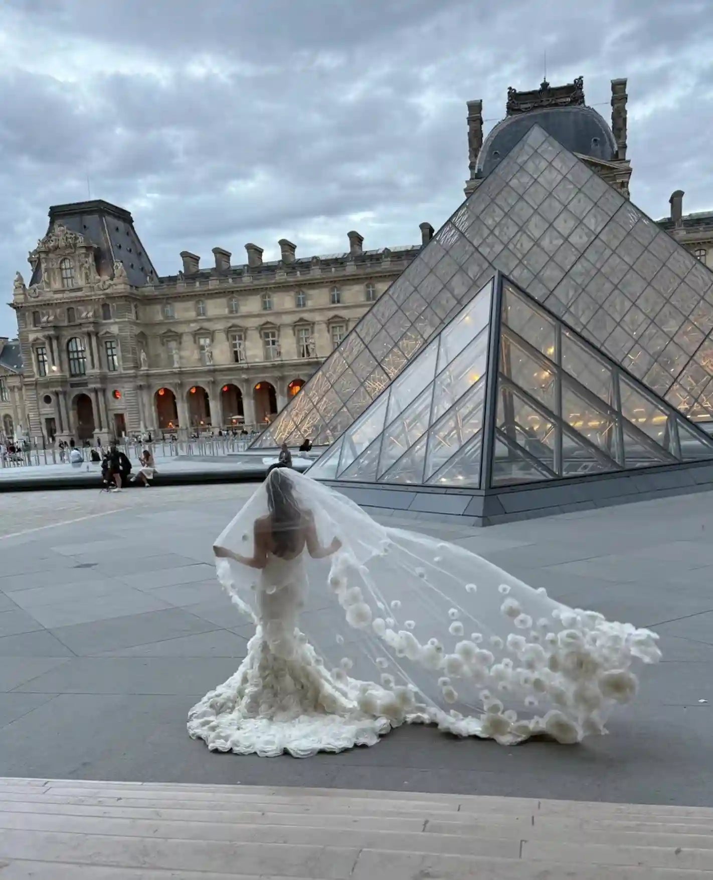 Mariée de dos devant la pyramide du Louvre portant un spectaculaire voile cathédrale orné de fleurs en relief 3D effet haute couture