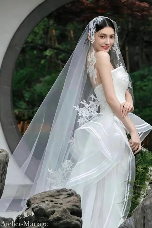 Vue d'ensemble d'une mariée portant un voile en dentelle brodée de 3 mètres avec une longue traîne, tenant un éventail, devant un jardin chinois à fenêtre ronde