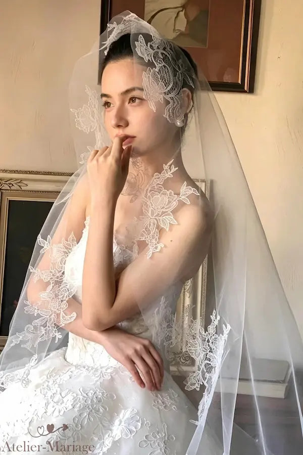 Pose rêveuse de la mariée, doigt sur les lèvres, portant le voile en dentelle fleurie de 3 mètres, élégance intemporelle