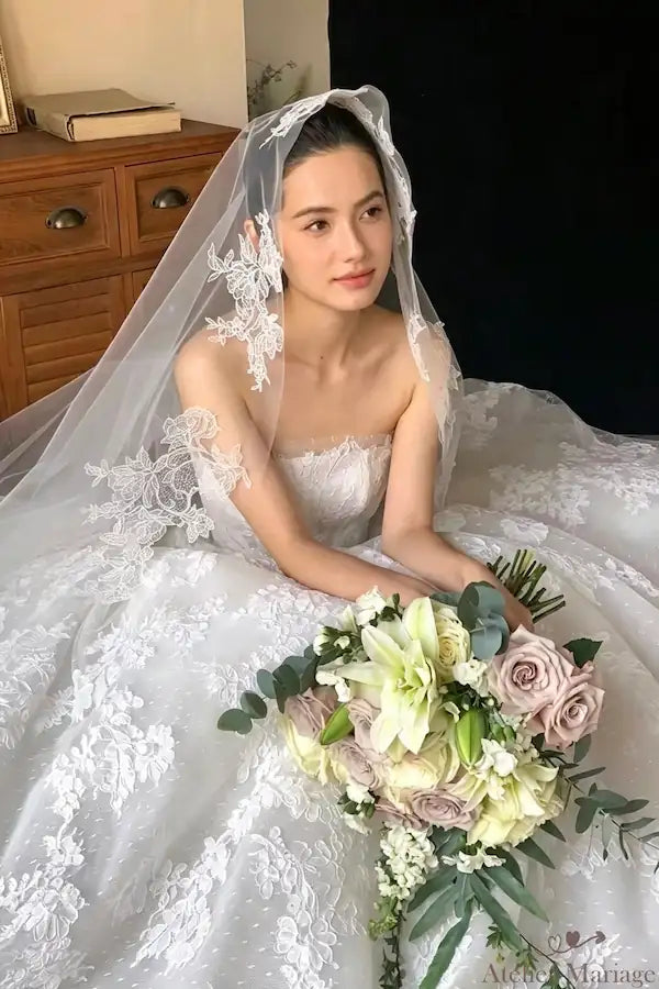 La mariée en robe bustier, tenant un bouquet de pivoines et roses, portant le voile en dentelle de 3 mètre